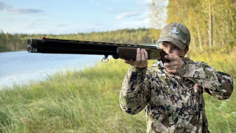 Silmälasipäinen nainen tähtää haulikolla.